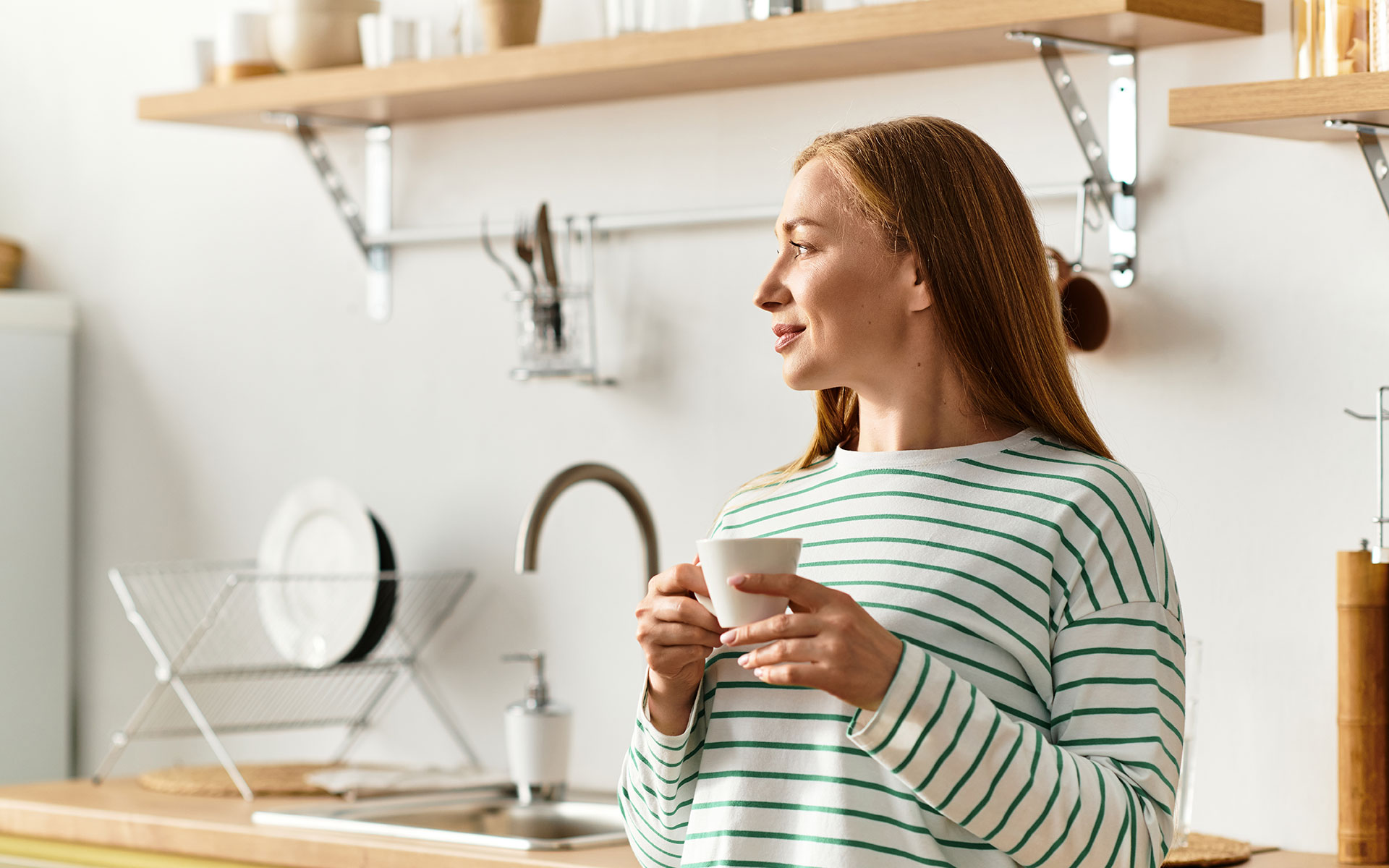 Ästhetische Behandlungen - Schönheitschirugie Bönen - Frau mit Kaffee steht in der Küche und schaut zum Fenster raus