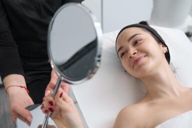 From above shot of unrecognizable  cosmetologist bringing a small mirror showing the results to a satisfied smiling female client lying on medical couch during beauty procedure in professional salon.